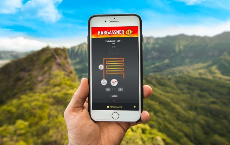 Smartphone in der Hand mit Hargassner Heizungsapp vor Landschaft