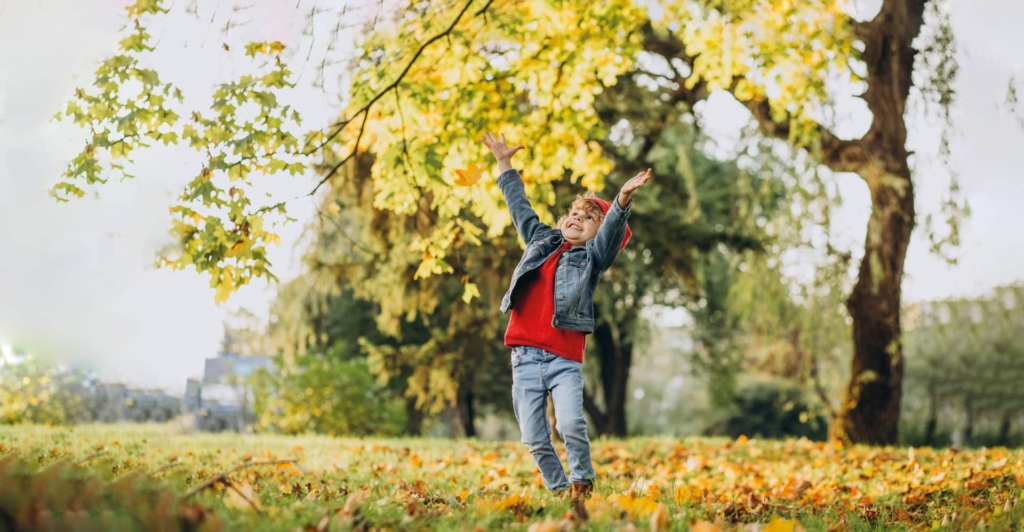 Kind lacht und streckt die Arme nach Oben zum herbstlichen Baum