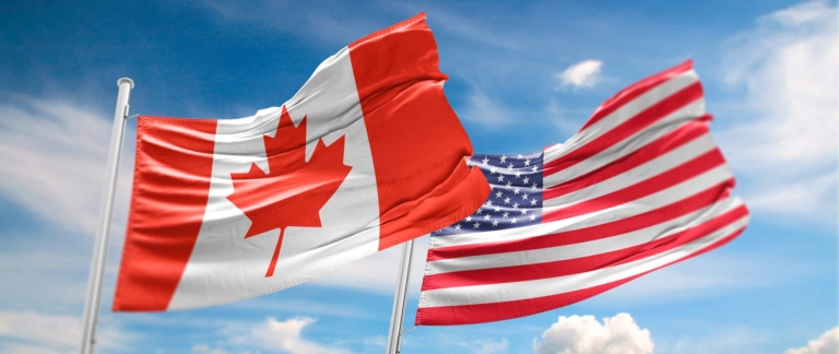 Drapeaux du Canada et des États-Unis devant un ciel bleu avec des nuages.