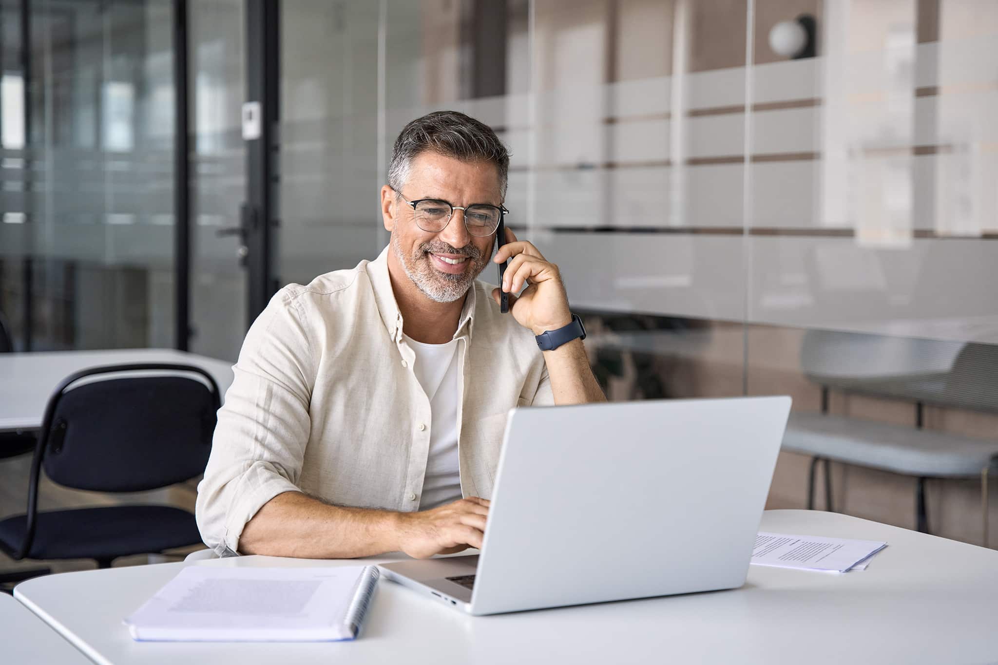 Man with glasses talks on the phone and looks into his laptop with a smile