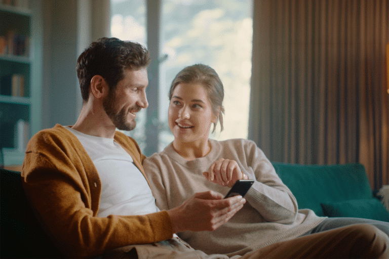 Paar sitzt auf Sofa im Wohnzimmer, Mann hält Handy in der Hand, sie blicken sich an und lächeln