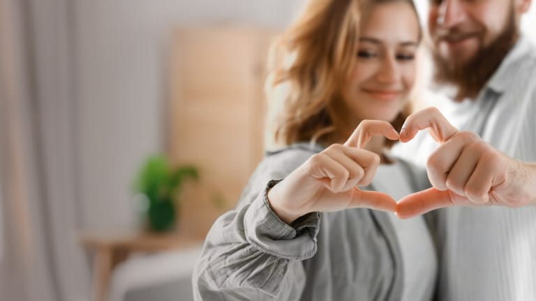 Mann und Frau formen mit ihren Händen Herzsymbol in einem hellen Wohnzimmer