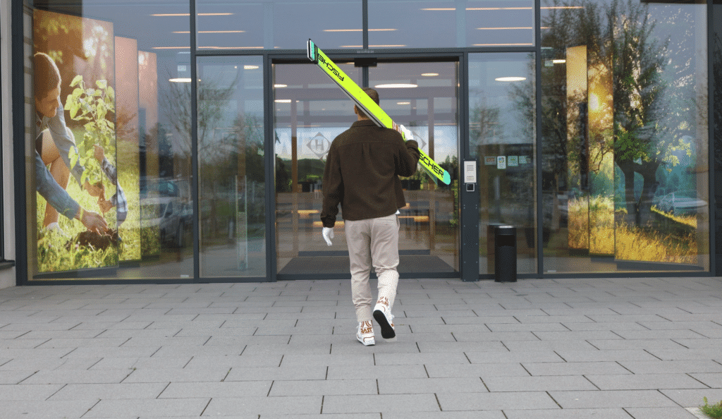 Daniel Huber mit angezogenen Skisprungschuhen, Handschuhen und Skisprungski geht in Richtung Eingang und Glasschiebetür