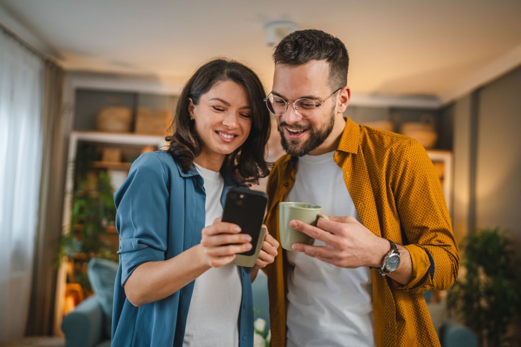 Junges, glückliches Paar im Wohnzimmer, sieht gemeinsam aufs Handy