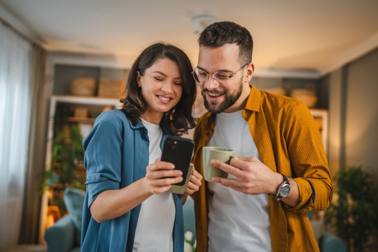 Junges, glückliches Paar im Wohnzimmer, sieht gemeinsam aufs Handy