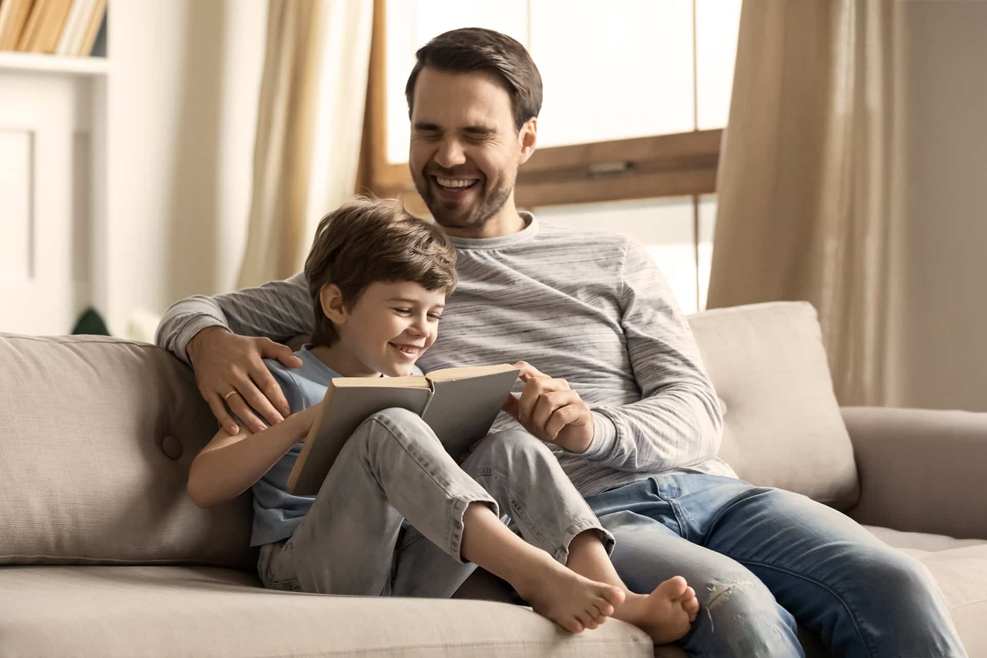 Mann und sein Sohn sitzen auf der Couch, lesen ein Buch und lachen